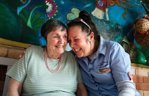 A Whiddon carer laughing together with a resident as she enjoys listening to music through her headphones.