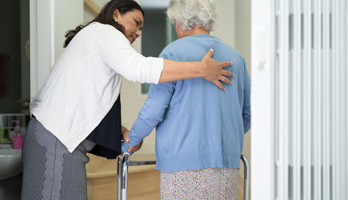 care worker helping older woman walk up stairs with mobility aid.
