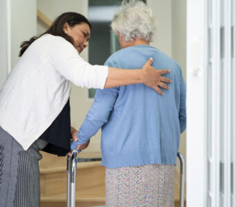 care worker helping older woman walk up stairs with mobility aid.